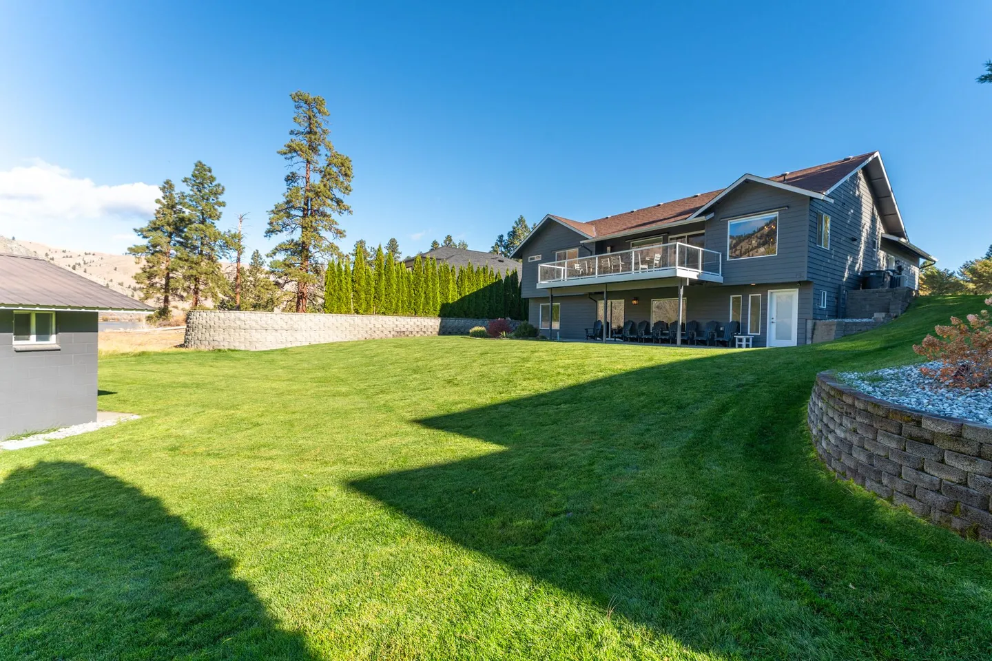 Exterior view of a multi-level gray house with a spacious green lawn, stone retaining walls, and an upper deck.