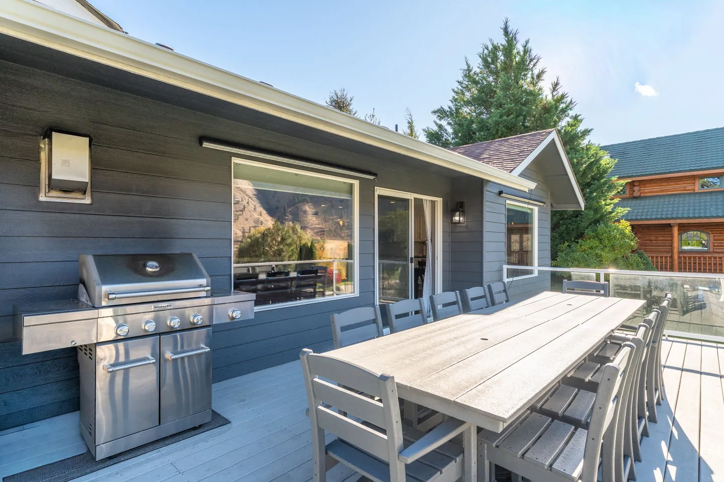 Outdoor deck featuring a stainless steel barbecue grill, a large dining table with multiple chairs, and glass railings under a clear sky.