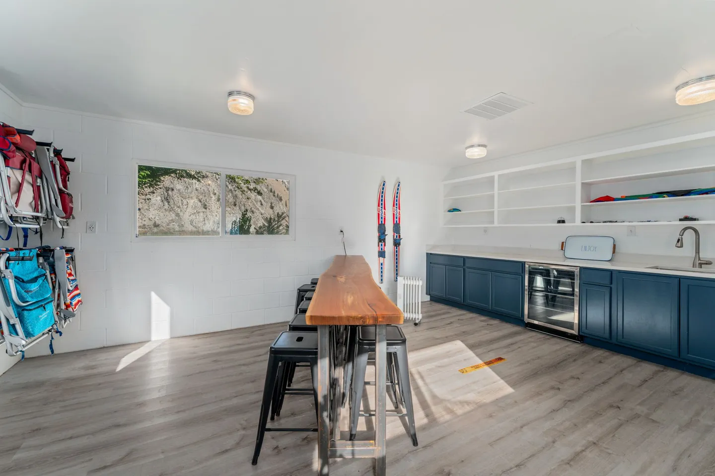 Bright recreation room featuring a long live-edge high-top table, navy wet bar cabinets, and beach chairs hung on a wall.