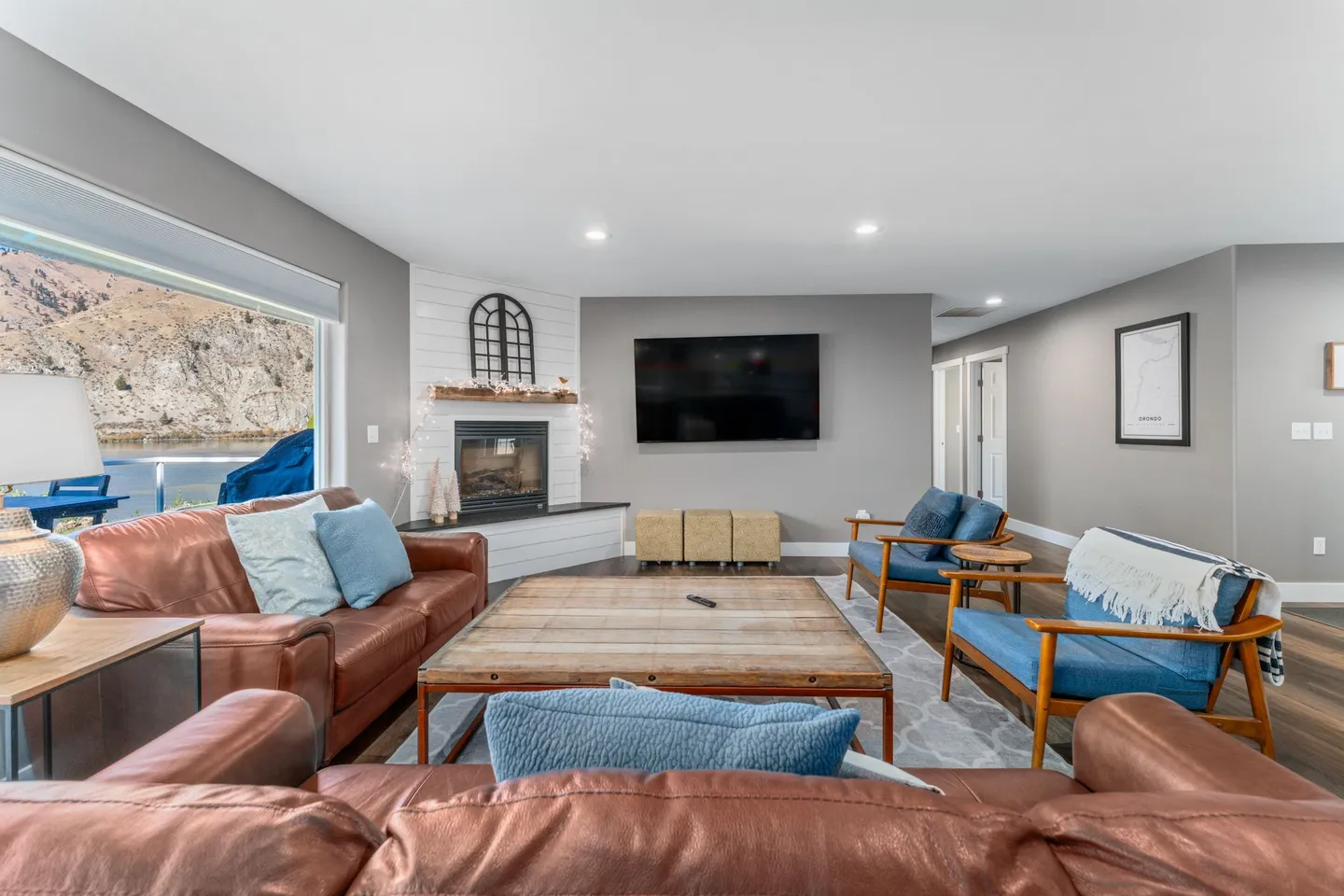 Living room with brown leather sofas, blue armchairs, a wood coffee table, a fireplace, and a wall-mounted TV with a view of water and hills outside.