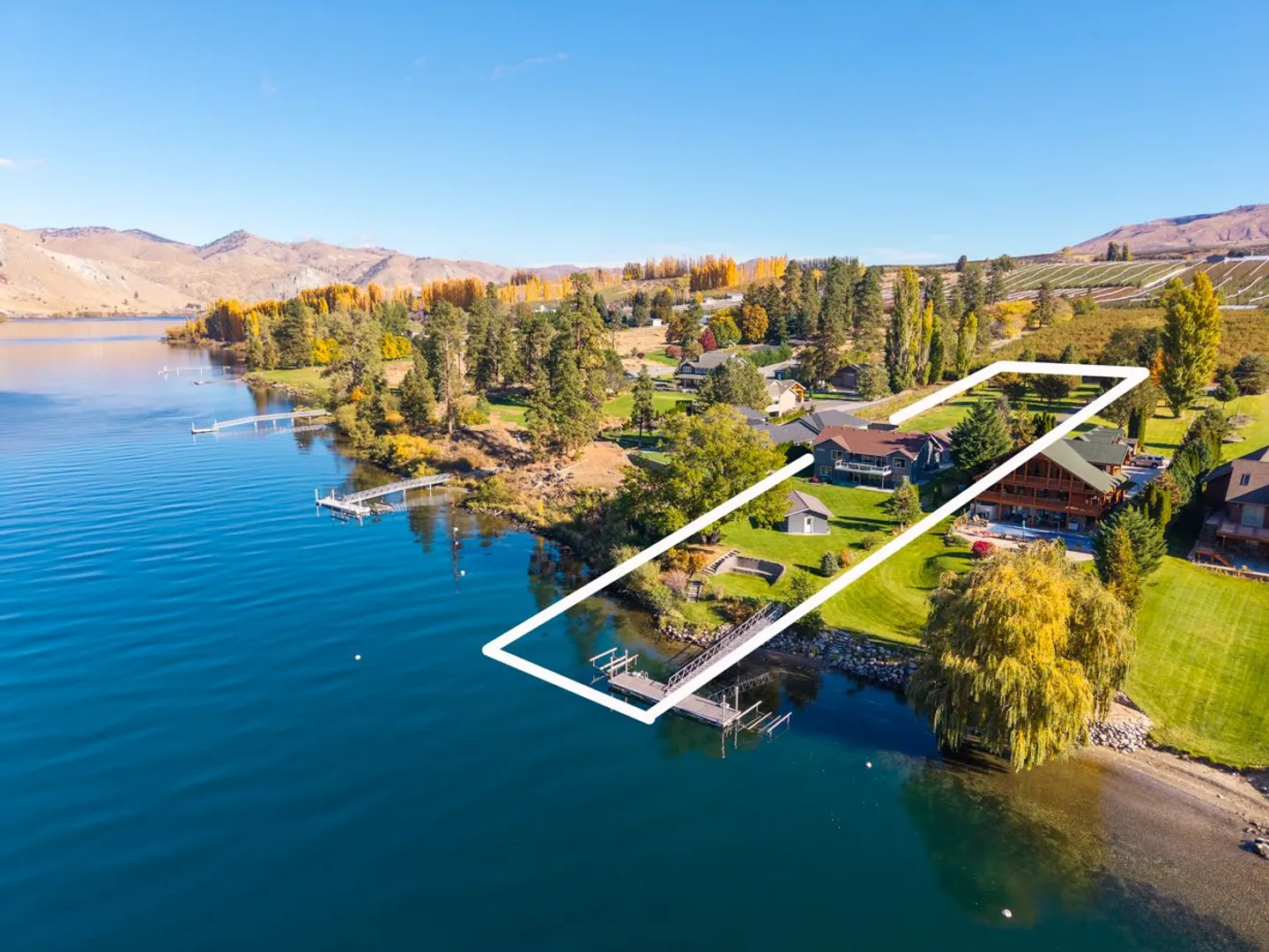 Aerial view of a lakefront property outlined in white with a private dock, green lawn, and surrounding hills.