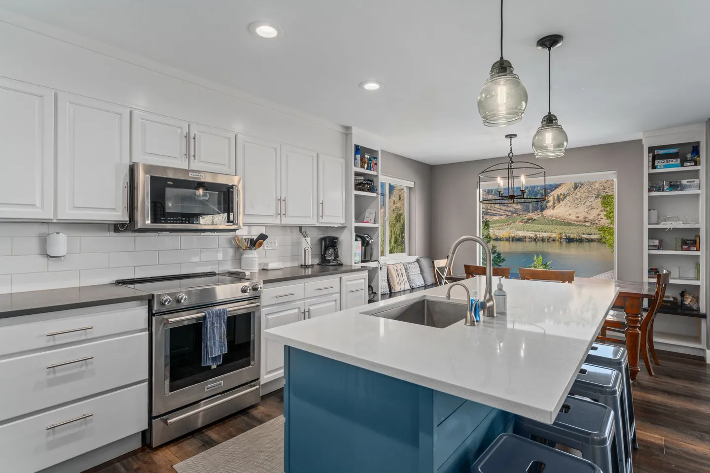Kitchen with white cabinetry, stainless steel appliances, and a blue island looking out toward a dining area and waterfront view through large windows.
