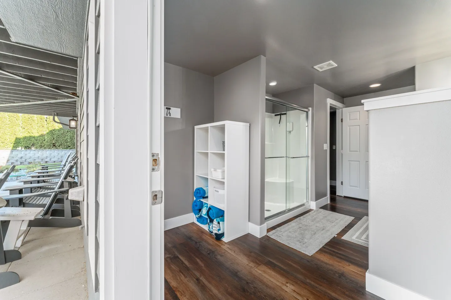 View from an open doorway into an indoor bathroom and changing area with dark wood-look floors, a white cubby shelf with towels, and a glass-enclosed shower.