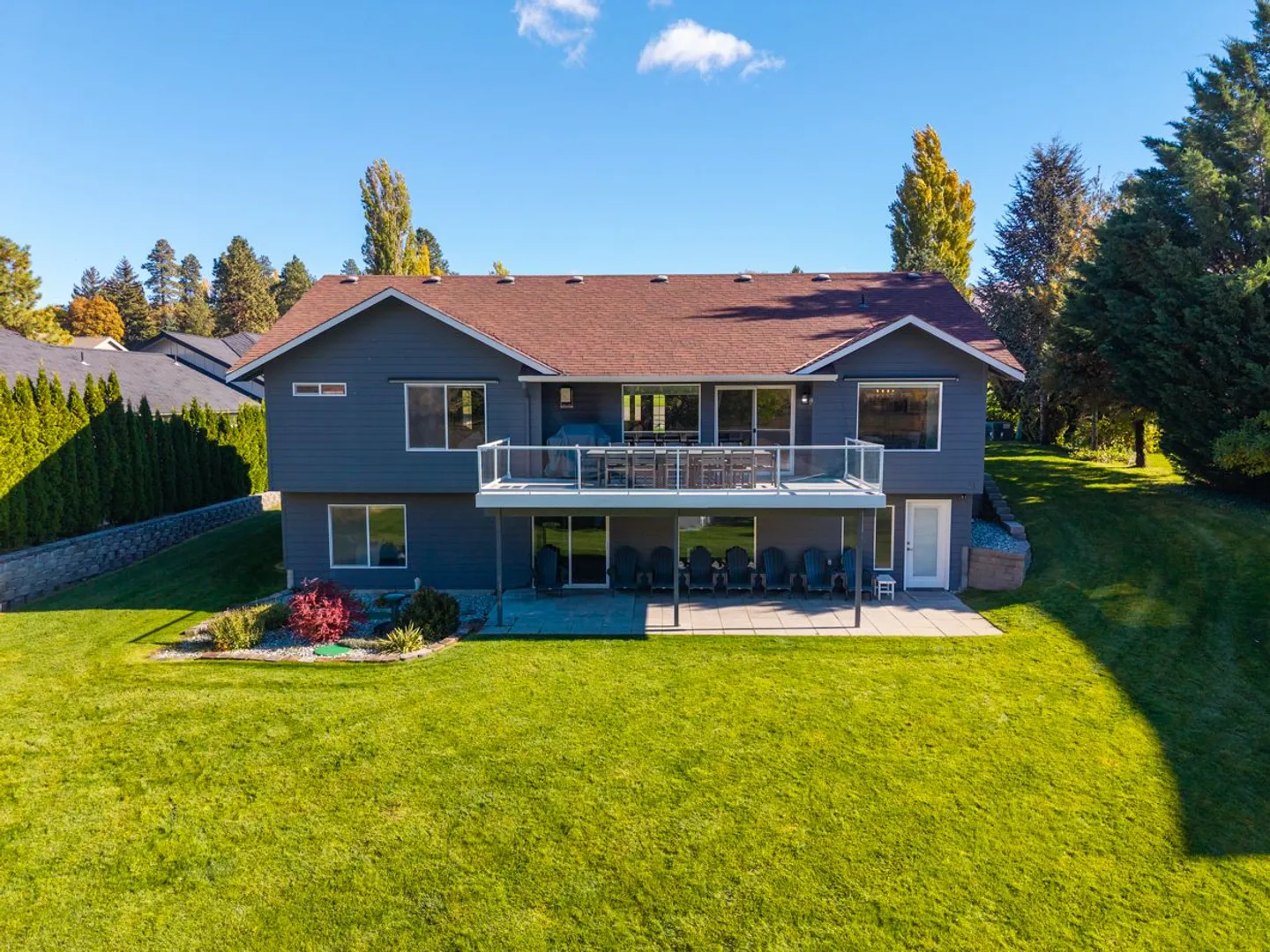 Rear exterior view of a two-story blue house featuring an upper-level deck, lower patio with seating, and a large green lawn.