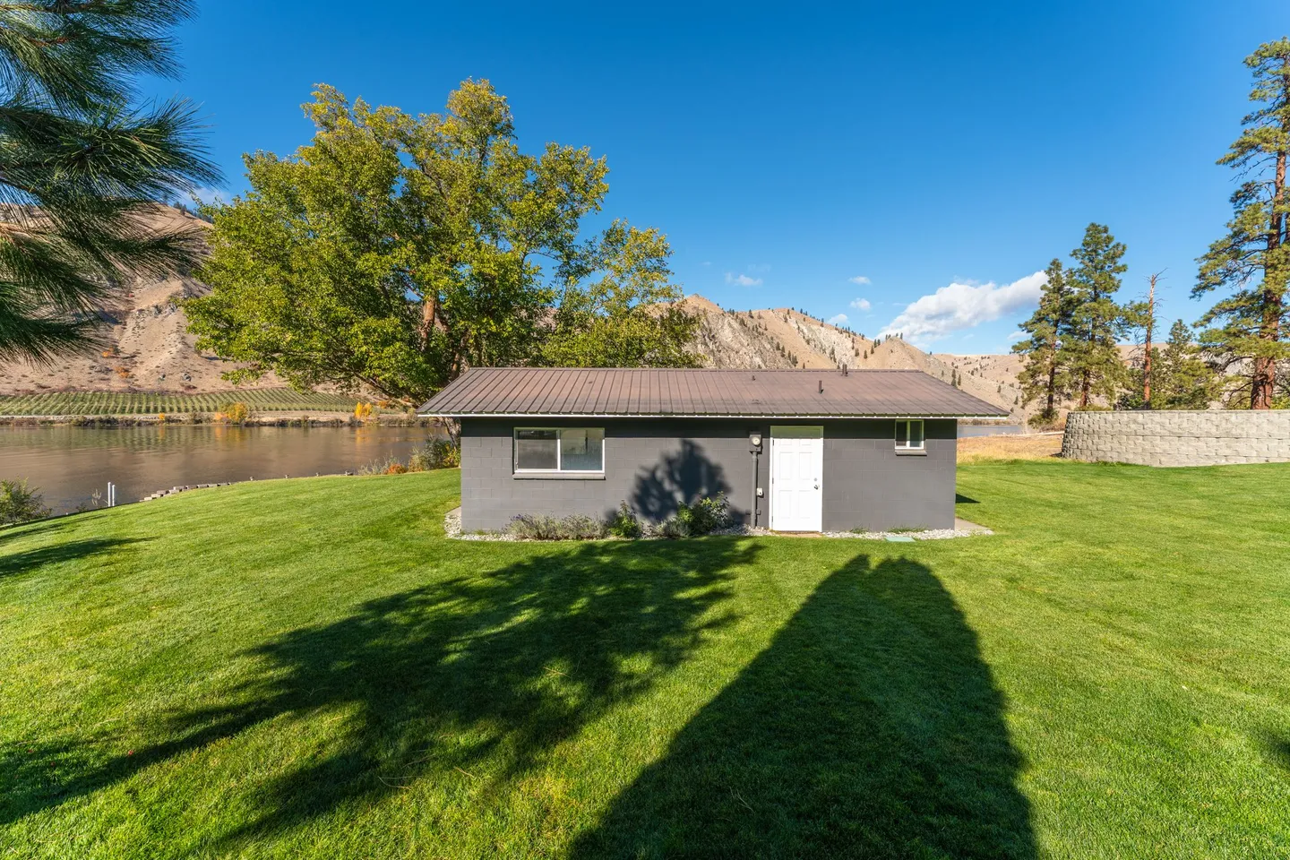 Exterior view of a small grey outbuilding on a green lawn beside a river with arid hills in the background.