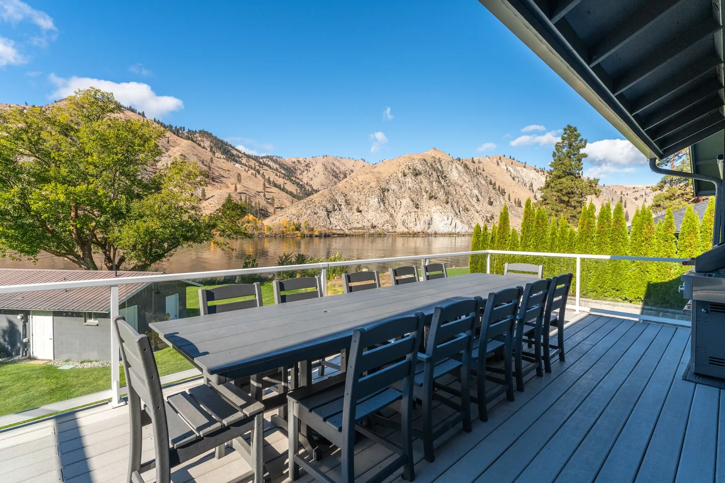Outdoor deck with a long dining table and chairs overlooking a wide river and rugged hills under a sunny blue sky.