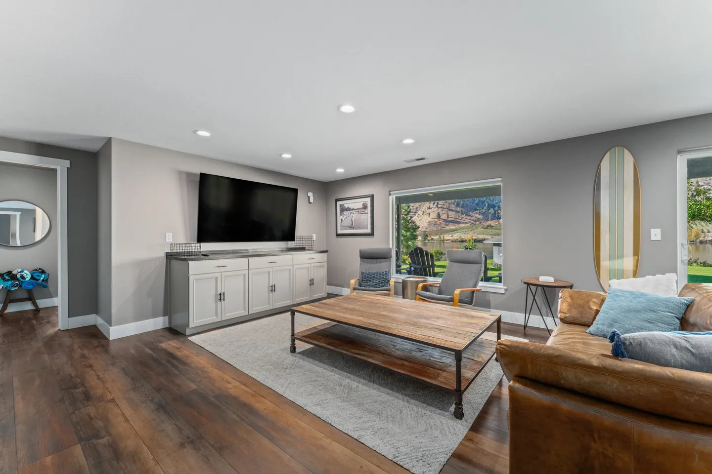 Living room with leather sofa, wooden coffee table, large wall-mounted TV over a sideboard cabinet, and window view of a mountain lake.