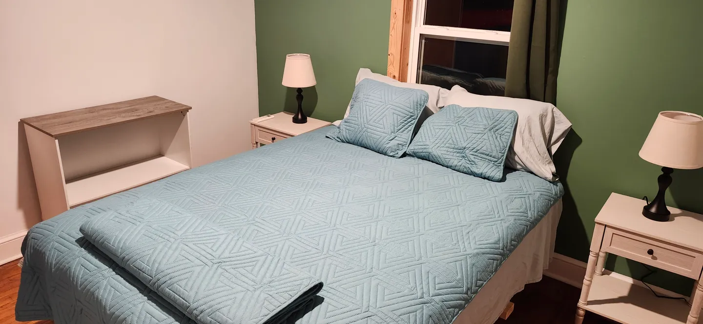 Bedroom with a bed made with a light blue patterned quilt, flanked by matching white nightstands with lamps against a green accent wall.