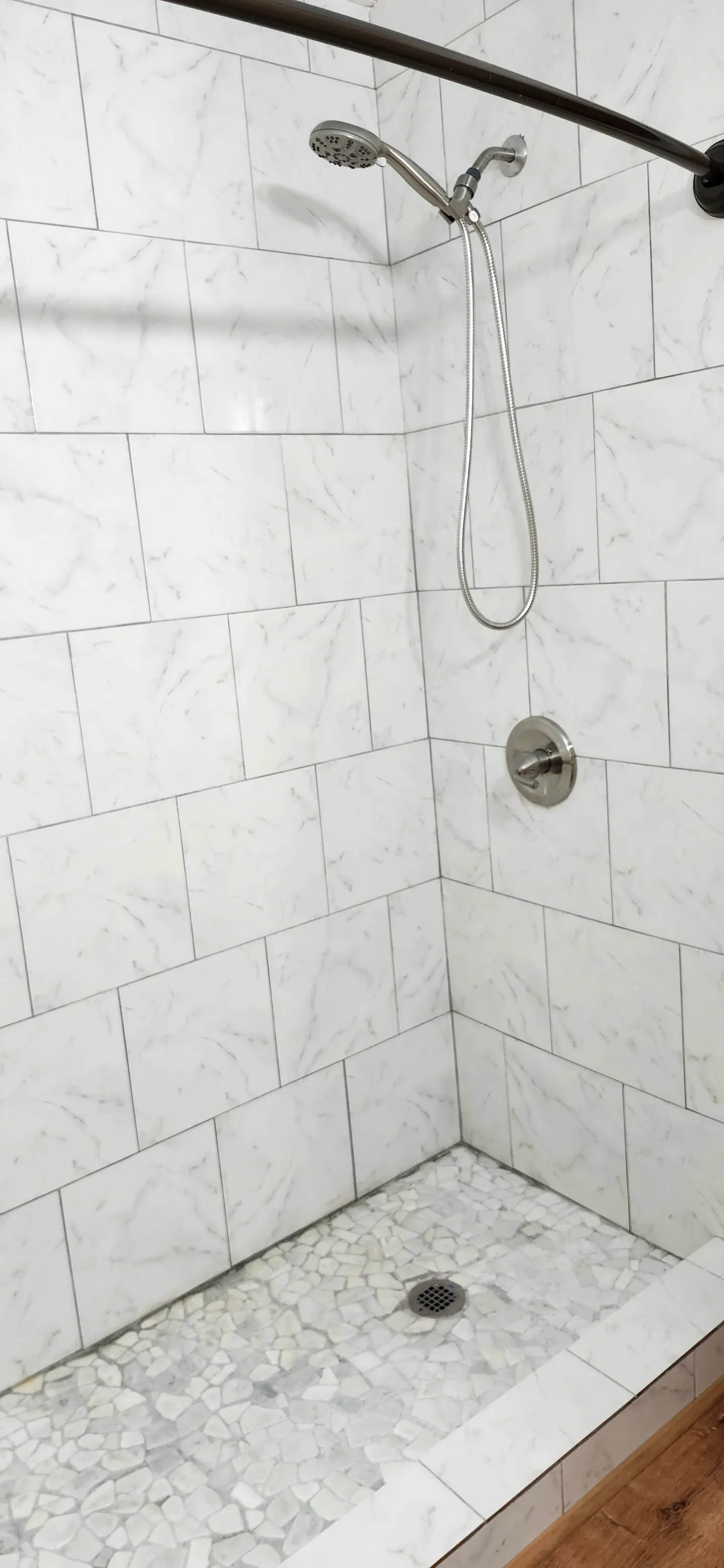 Walk-in shower tiled in white marble-look subway tiles with a stone pebble floor, brushed metal showerhead, and curtain rod.
