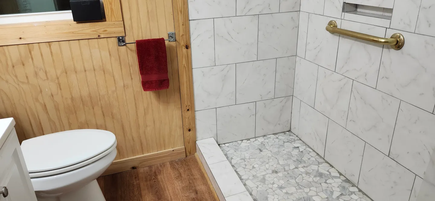 Bathroom showing a walk-in shower with marble-look wall tiles, pebble tile floor, a gold grab bar, next to wood-paneled walls and a toilet.