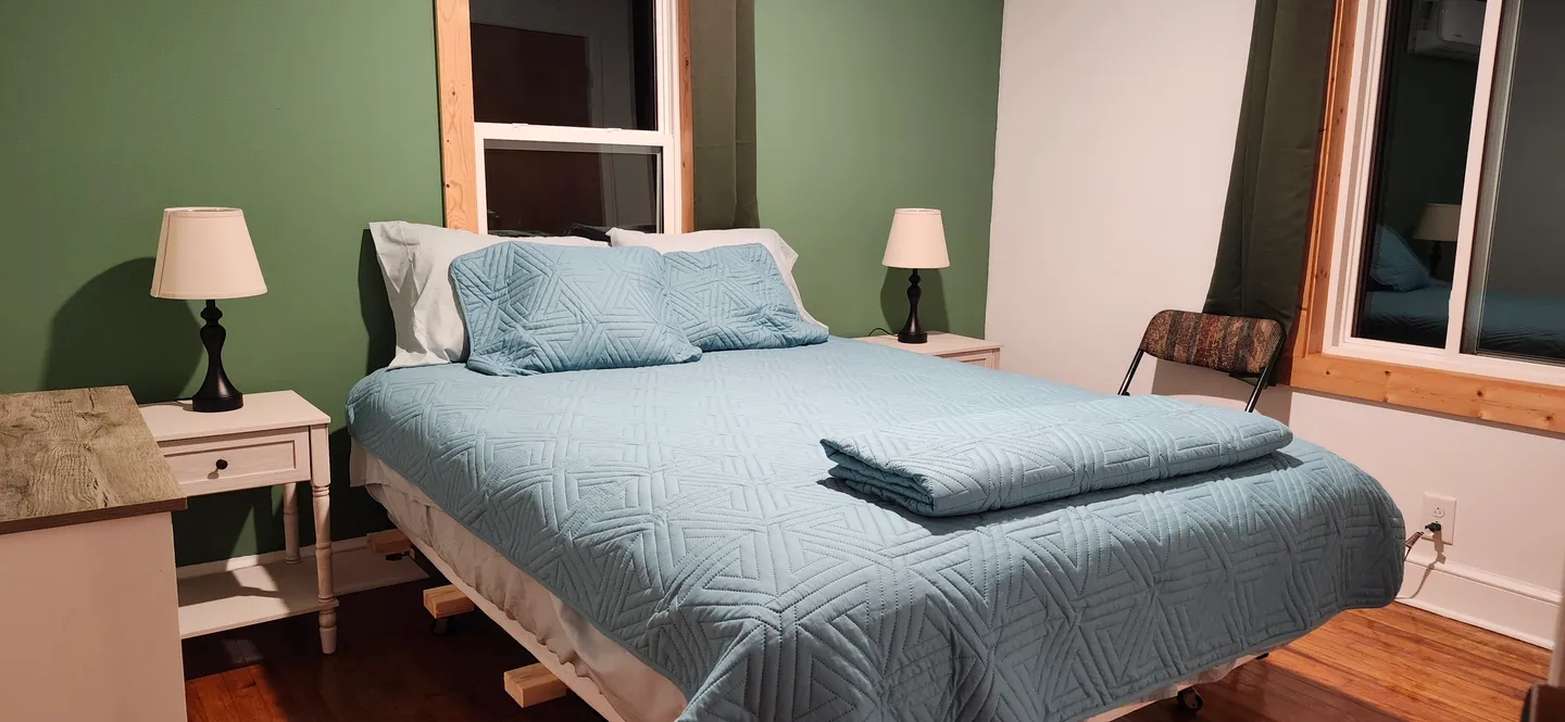 Bedroom with a bed made with a light blue textured quilt, flanked by nightstands with matching lamps against a green accent wall.