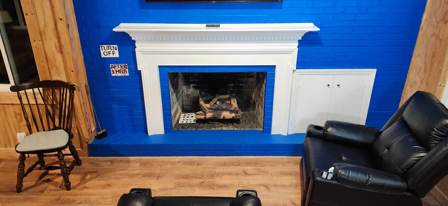 A painted bright blue brick fireplace wall with a white mantel and gas logs, flanked by a wooden chair and leather recliner.