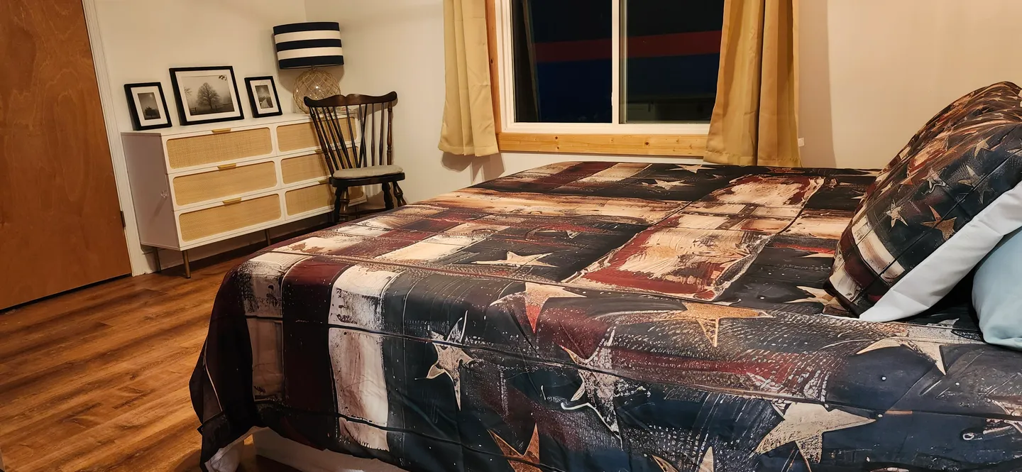 Bedroom featuring a bed with a star-patterned comforter, a white and woven dresser, and a window with gold curtains.