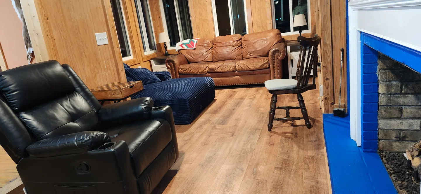Living room with wood-paneled walls, leather seating, a wood-look floor, and a fireplace with a blue-painted brick hearth.