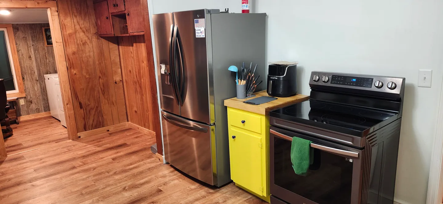 Kitchen area featuring a stainless steel refrigerator, an electric range, and a small yellow cabinet, with wood-paneled walls and an adjacent laundry room visible in the background.