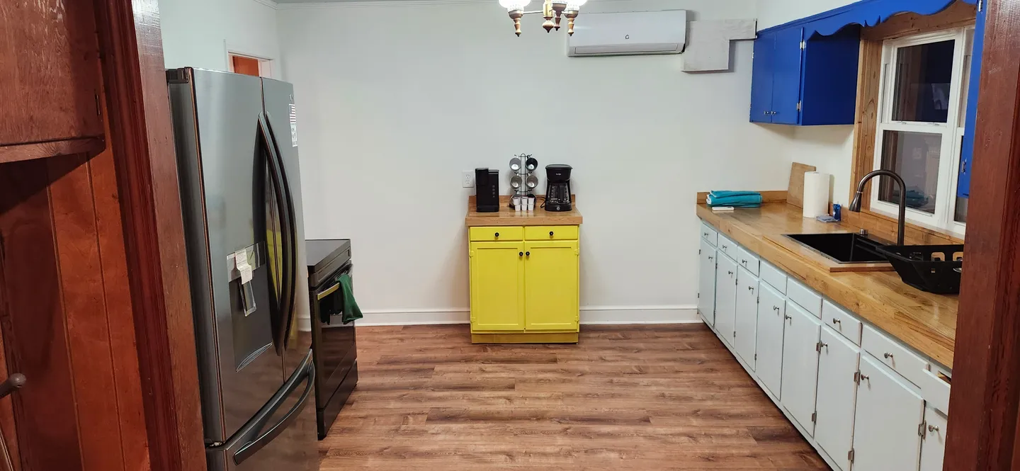 Kitchen with stainless steel refrigerator, white and bright blue cabinets, a yellow standalone coffee station, and butcher block countertops.