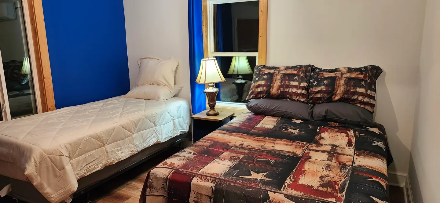 Bedroom with two beds, a nightstand with a lamp between them, a blue accent wall, and a window.