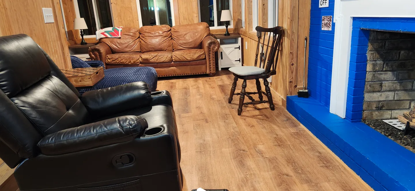 Living area featuring wood-paneled walls, wood-look flooring, a vibrant blue painted brick fireplace, a black recliner, and a brown leather sofa.