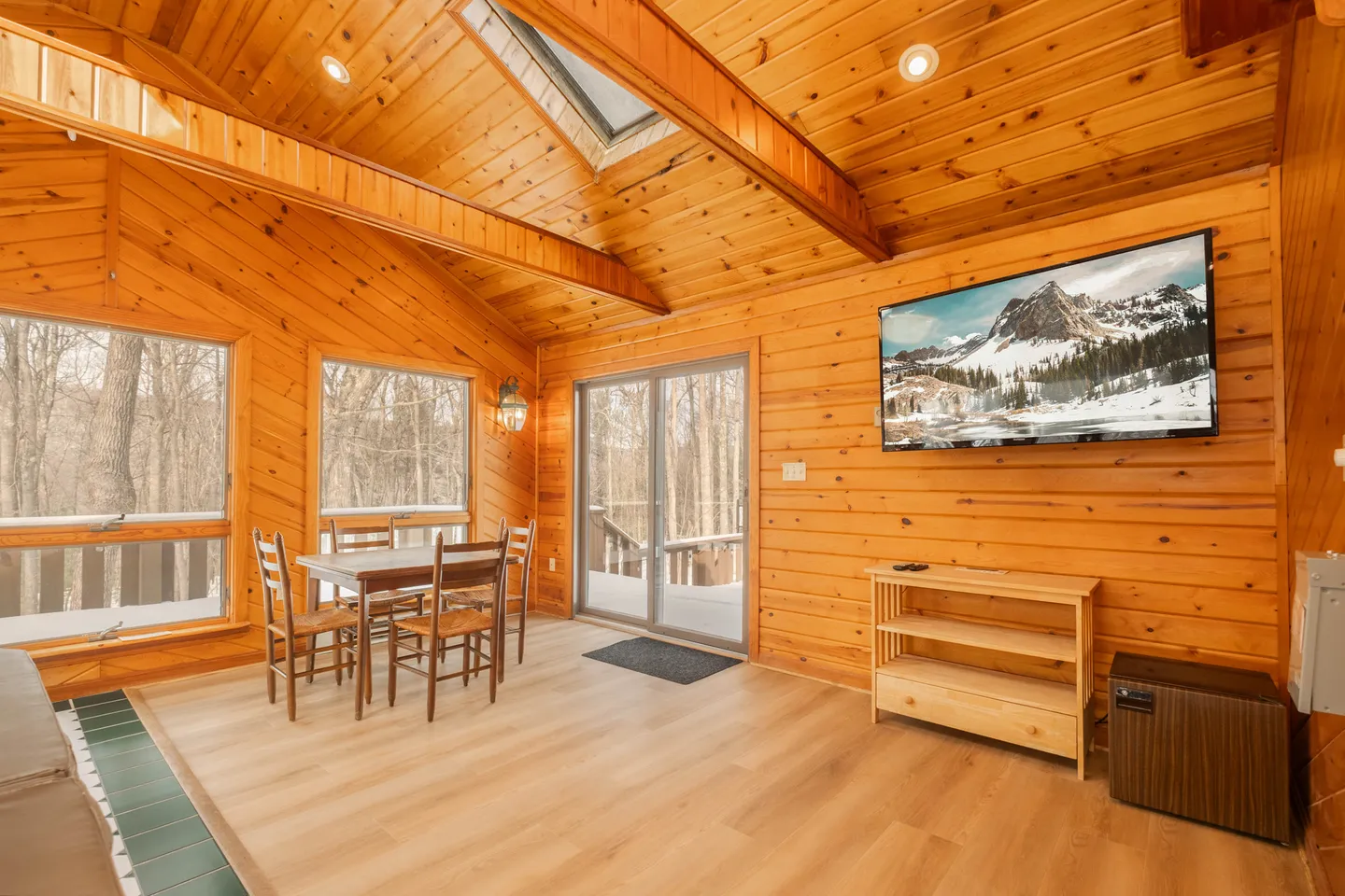 Sunroom with wood-paneled walls and ceiling, dining table with chairs, mounted television, and sliding glass door leading outside.