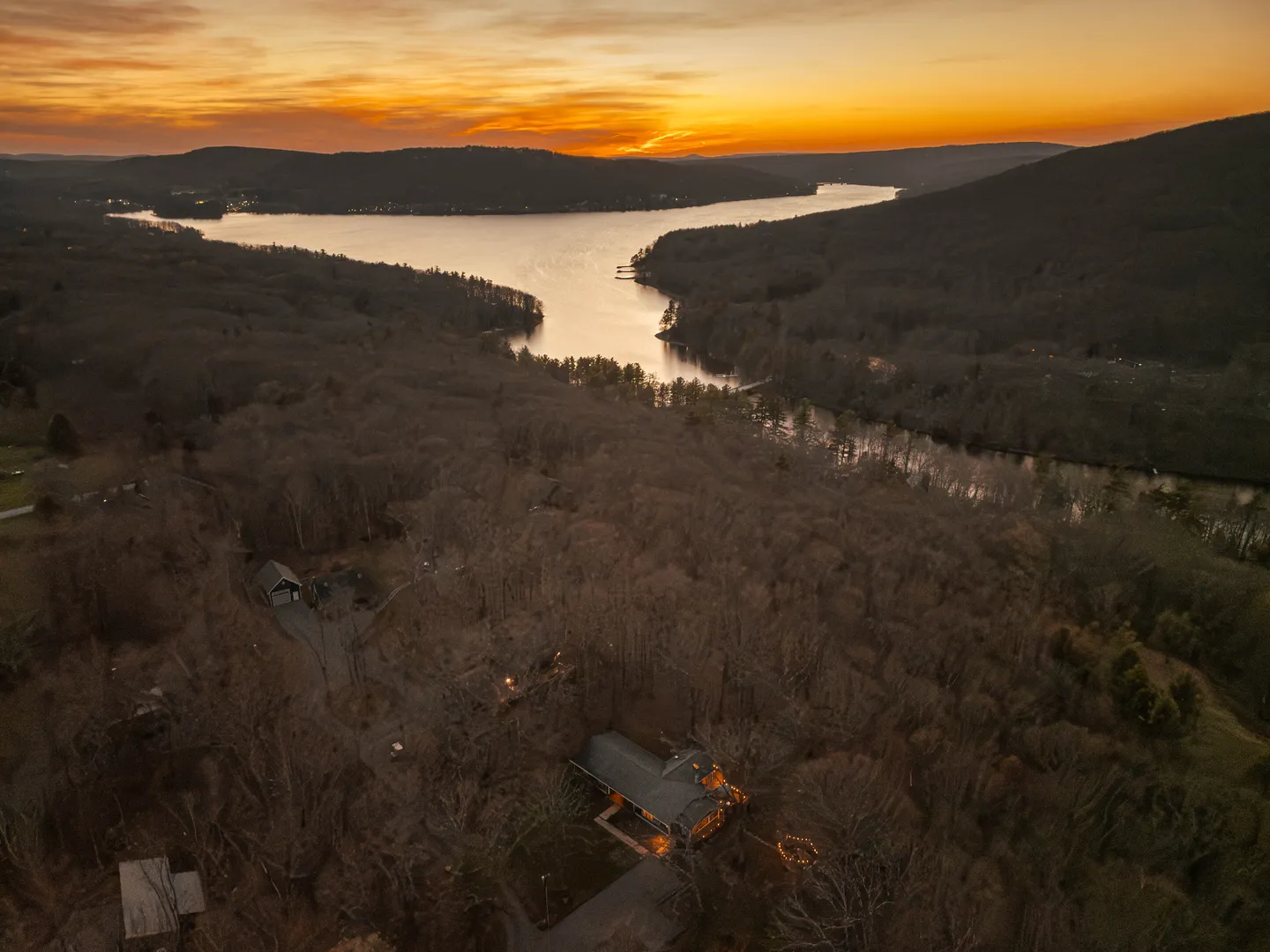 Aerial sunset view of a wooded property with glowing lights overlooking a river or lake framed by rolling hills.
