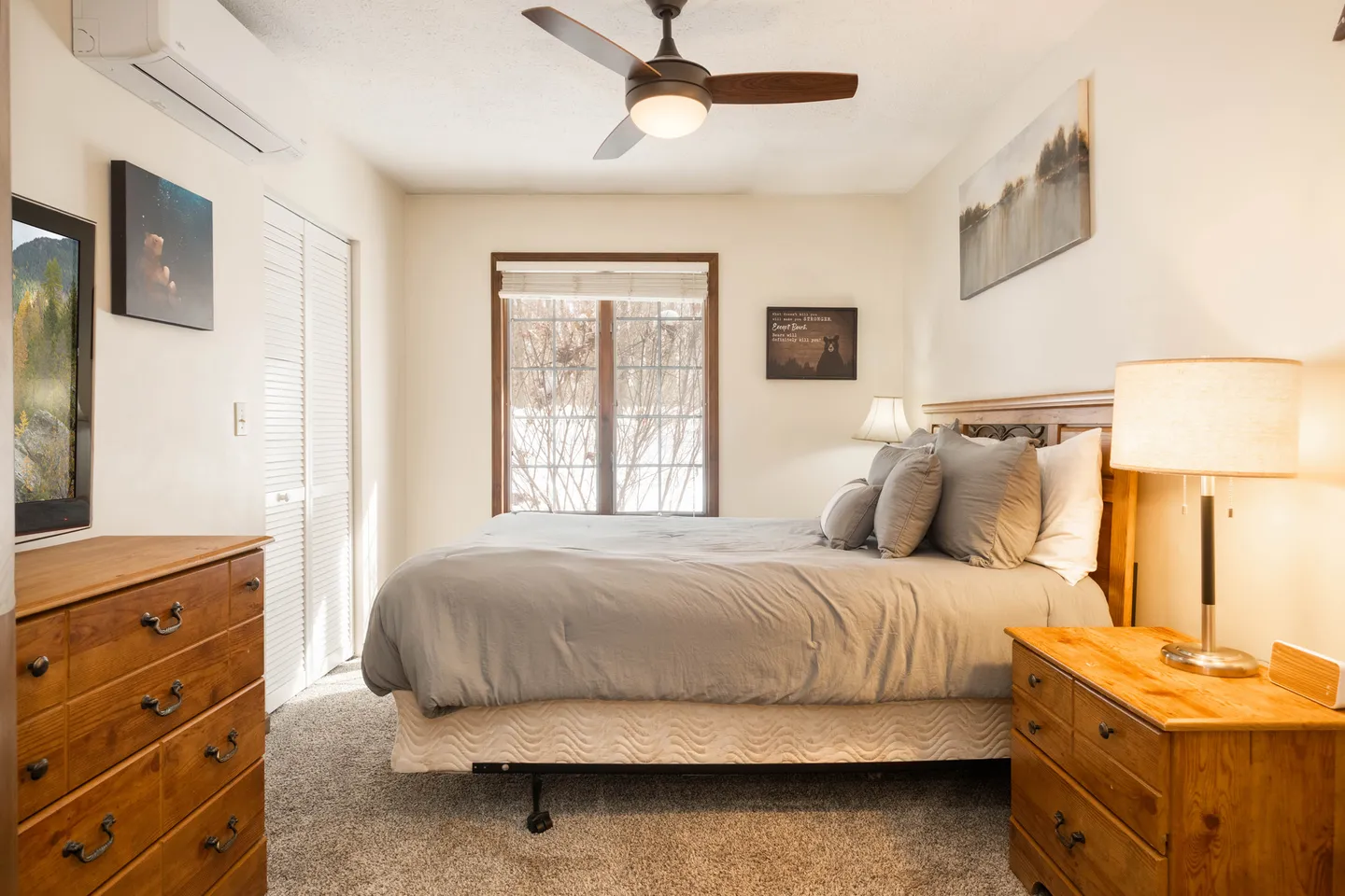 Bedroom with a bed made up with grey bedding, wooden dressers, a ceiling fan, a window, and carpet flooring.