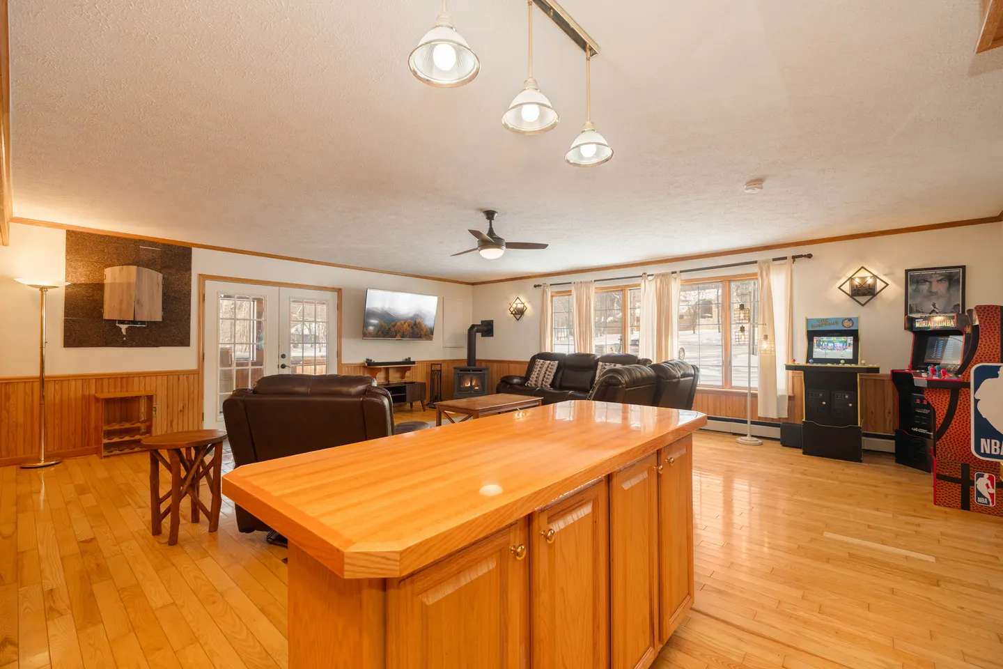 Open living area with wood floors, kitchen counter in foreground, leather seating, a wall-mounted TV, wood stove, and arcade games.