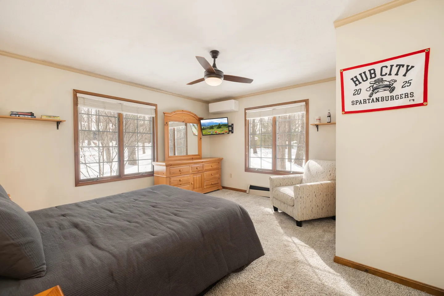 Bedroom featuring a bed with a gray coverlet, carpeted floor, ceiling fan, wooden dresser, armchair, and two windows showing snow outside.