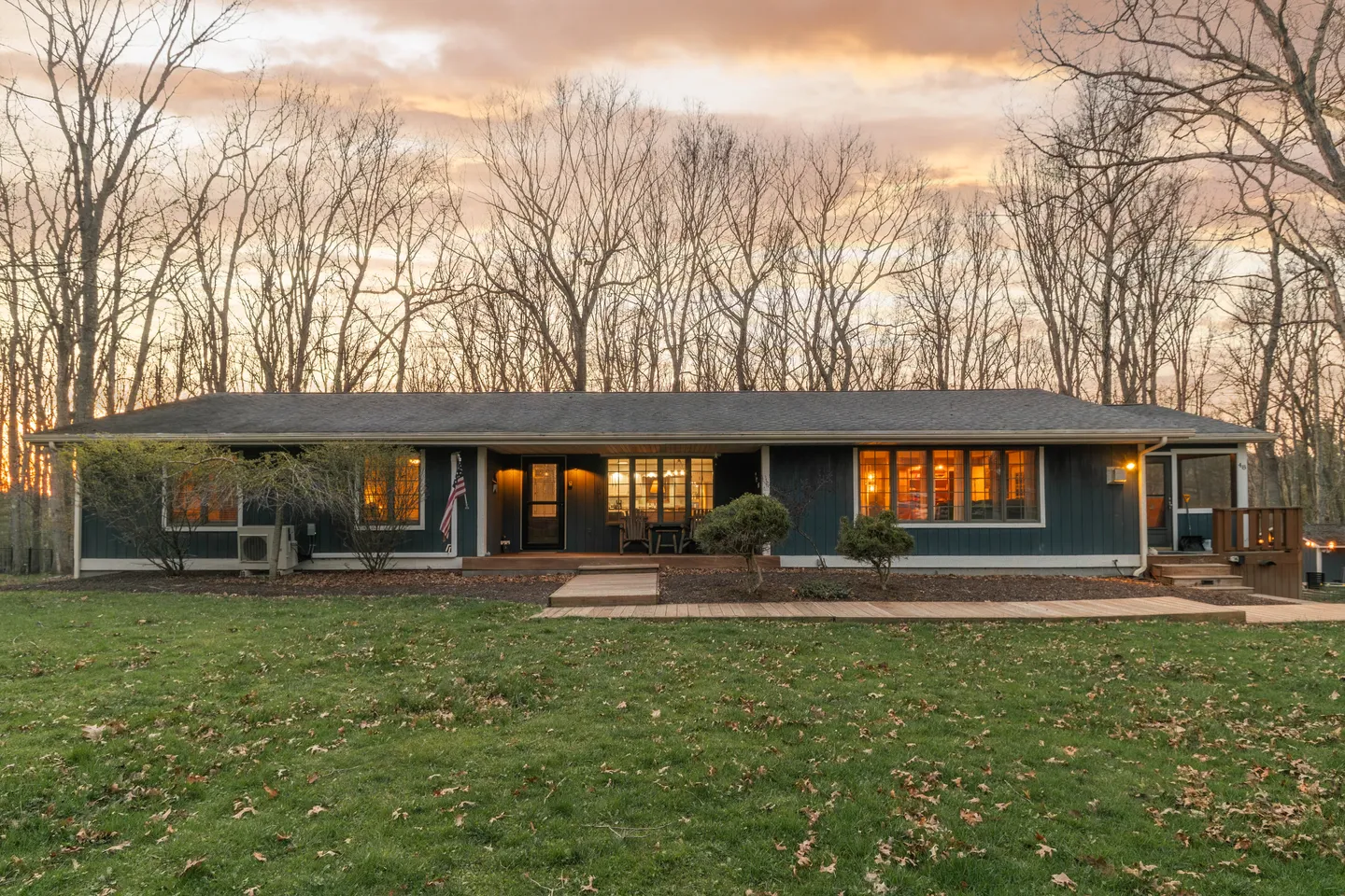 Exterior dusk view of a single-story dark blue house with warm interior lights and a grassy yard surrounded by trees.