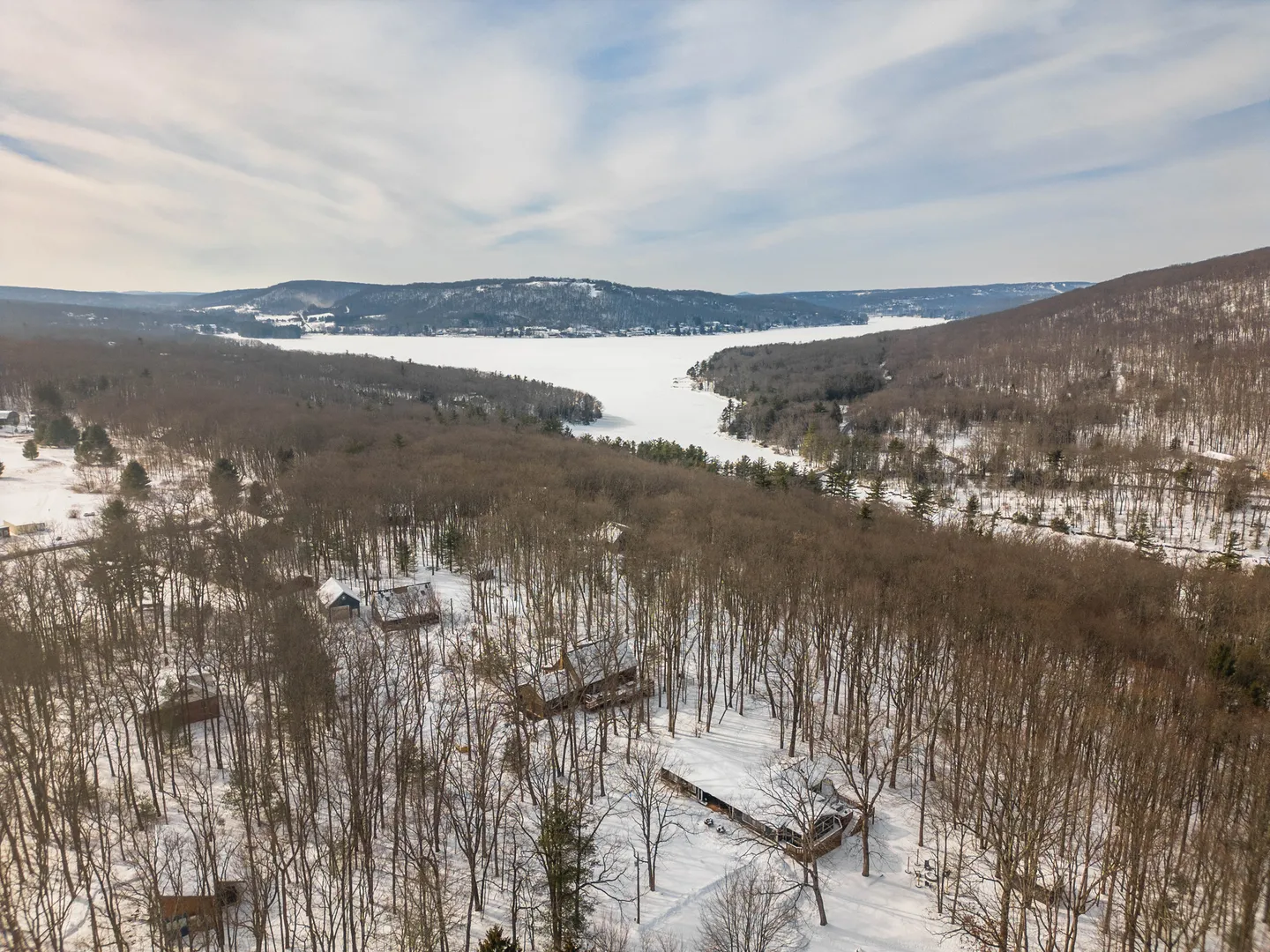 Aerial view of cabins nestled in a snow-covered forest overlooking a frozen lake and surrounding hills under a partly cloudy sky.