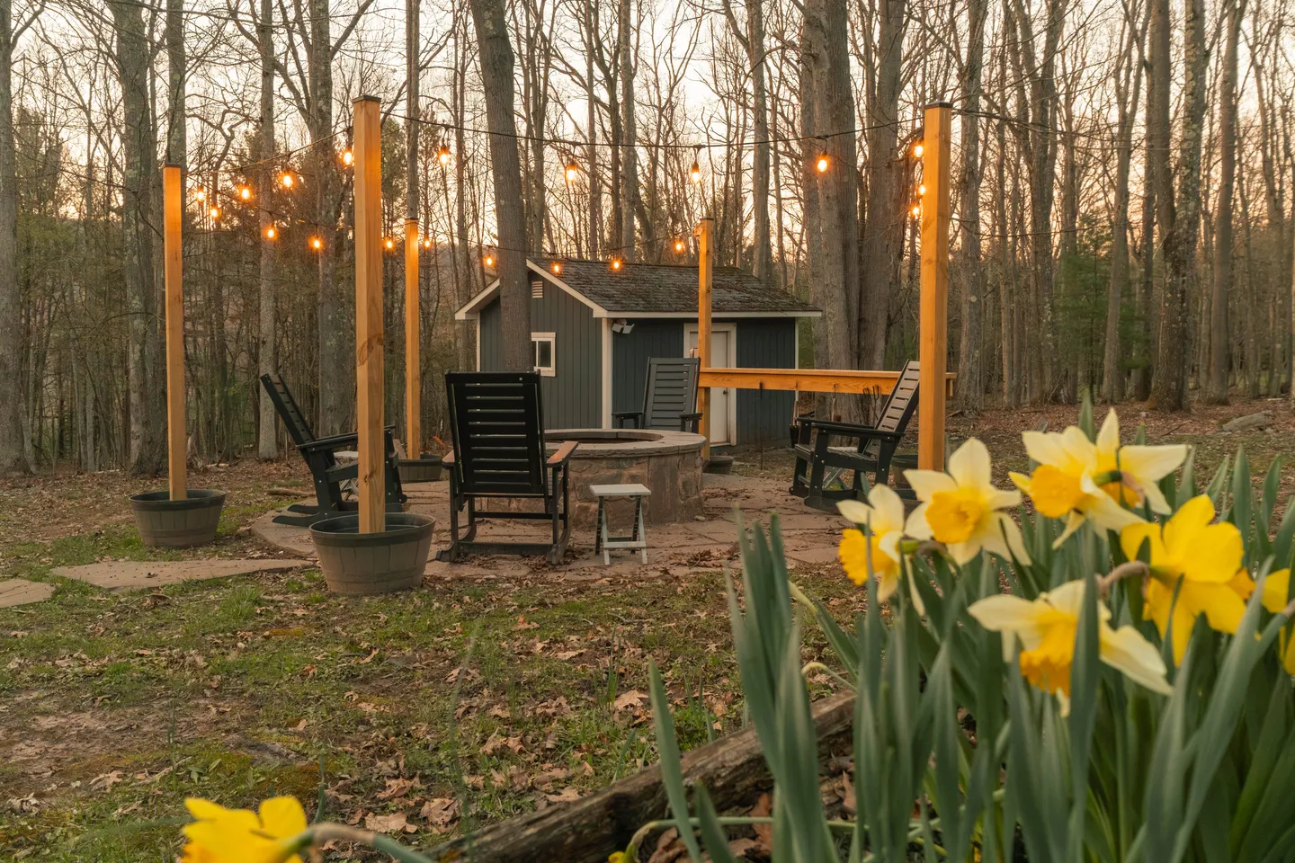 Outdoor fire pit area with rocking chairs and string lights surrounded by woods and yellow daffodils in the foreground.