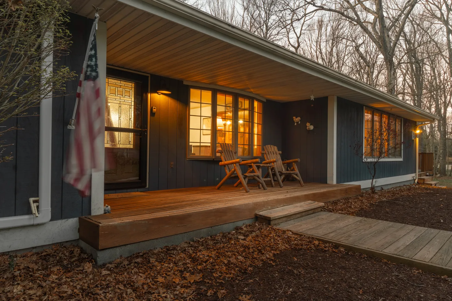 Front porch of a house at dusk with wooden deck, two armchairs, large bay window, and exterior lights glowing.