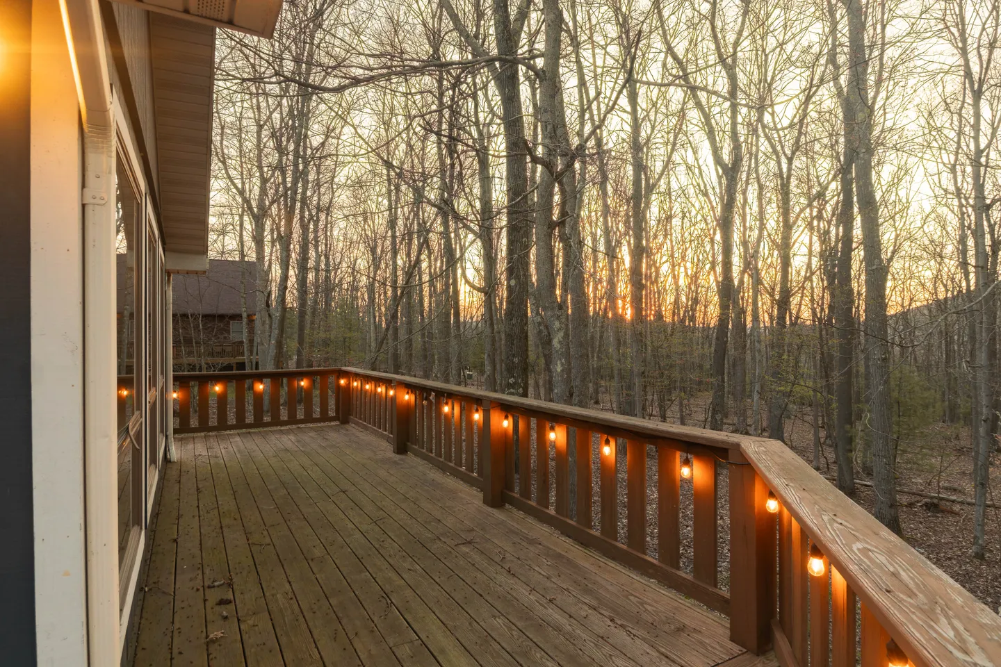 Wooden deck lined with string lights along the railing, overlooking bare trees at sunset.