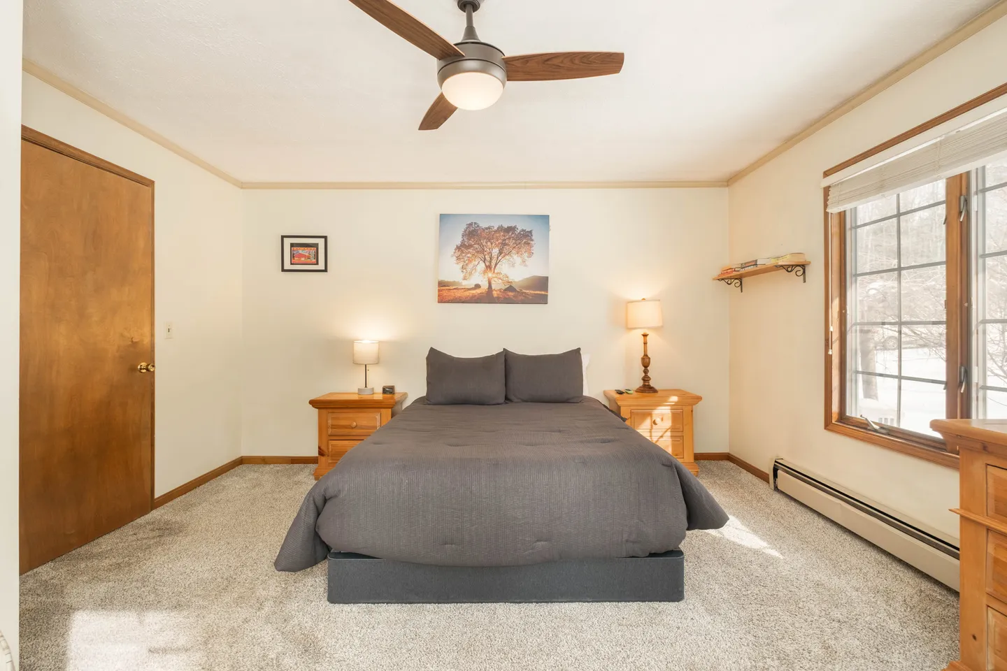 Bedroom with a made bed flanked by wooden nightstands, carpeted floor, ceiling fan, and window.