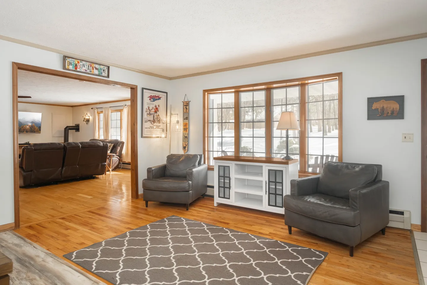 Living area with wood-look flooring, two grey armchairs, a patterned area rug, and a large multi-pane window looking out at snow.