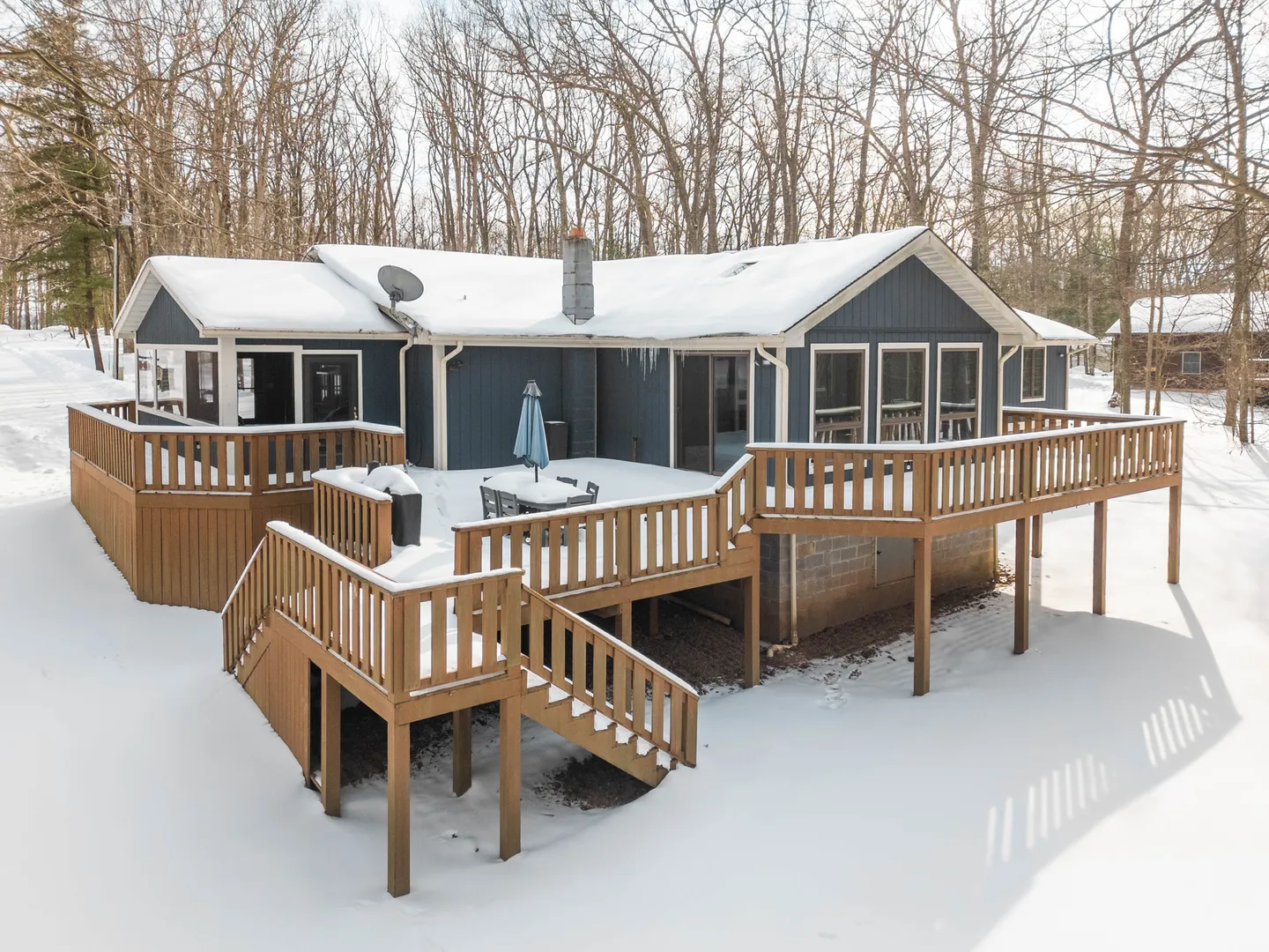 Exterior rear view of a blue house with a multi-level wooden deck surrounded by snow and winter trees.