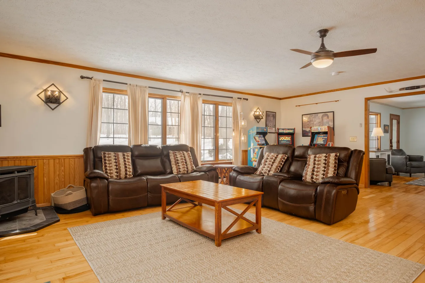 Living room with brown reclining sofas, a wooden coffee table on an area rug, a wood stove, and classic arcade machines in the background.