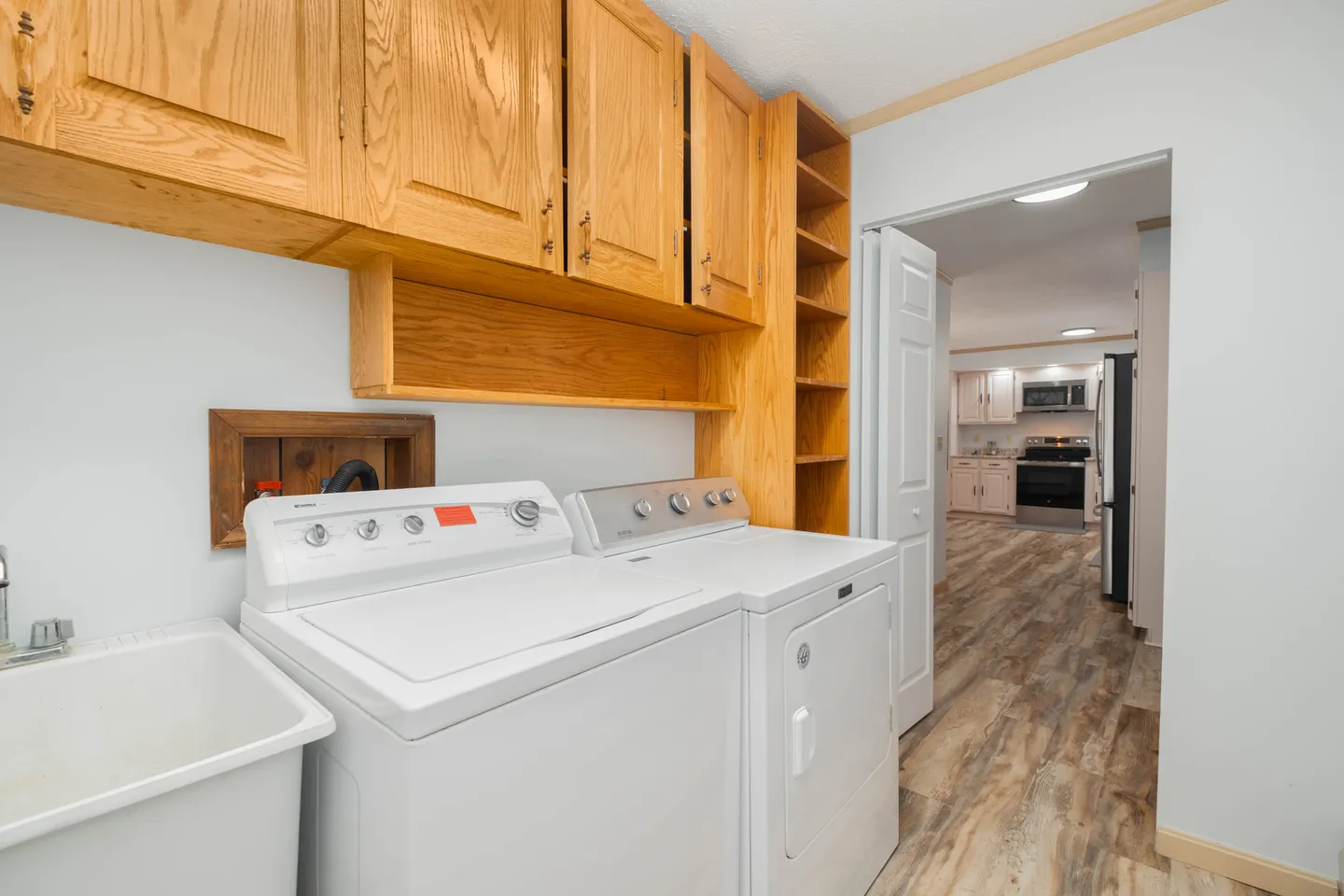 A laundry room with a white washing machine, dryer, utility sink, and light wood overhead cabinets looking into the kitchen.