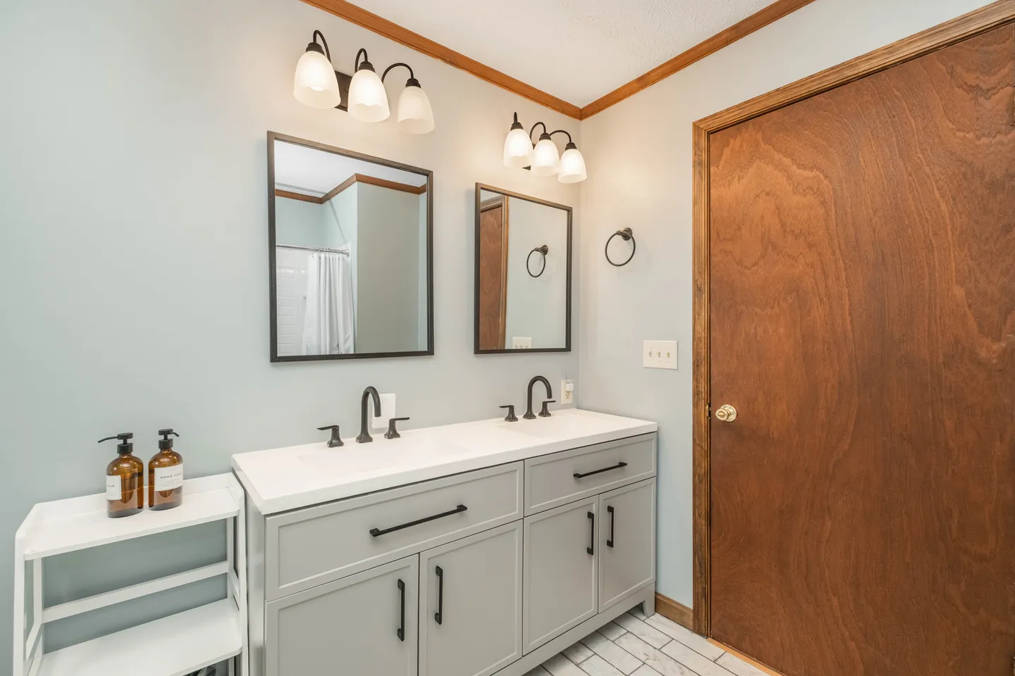 Bathroom vanity with two sinks, dark faucets, dual mirrors with overhead light fixtures, and a wooden door.