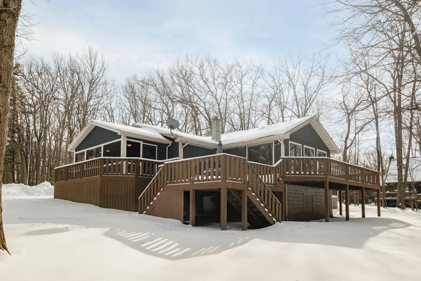 Rear exterior of a dark gray home with a large wooden deck and staircase surrounded by snow-covered ground and bare trees.