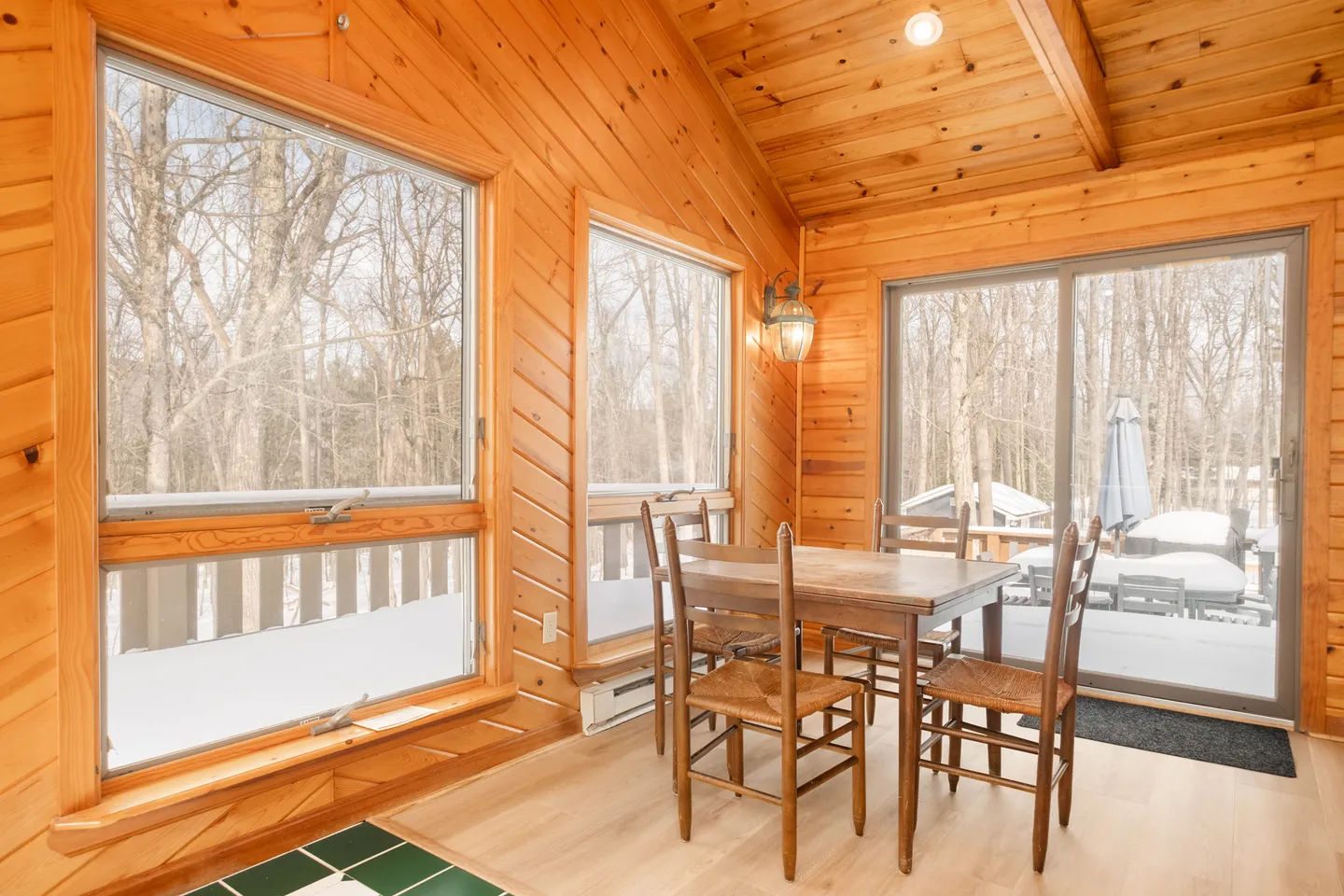 Sunroom dining area with wood-paneled walls and ceiling, dining table with four chairs, and large windows looking out onto a snowy deck and woods.