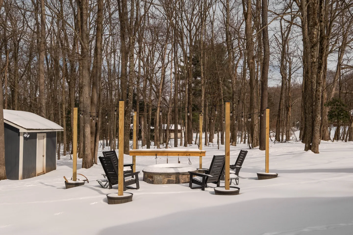 Snow-covered backyard featuring a circular stone fire pit surrounded by Adirondack chairs, string lights, and a small shed in front of bare trees.