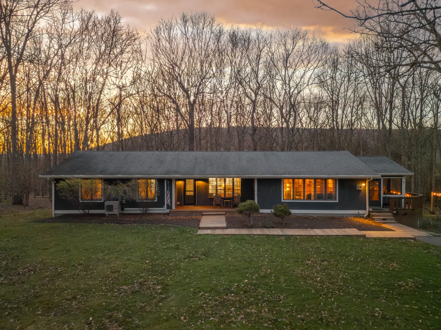 Exterior dusk view of a single-story dark-sided ranch home surrounded by woods with illuminated windows.