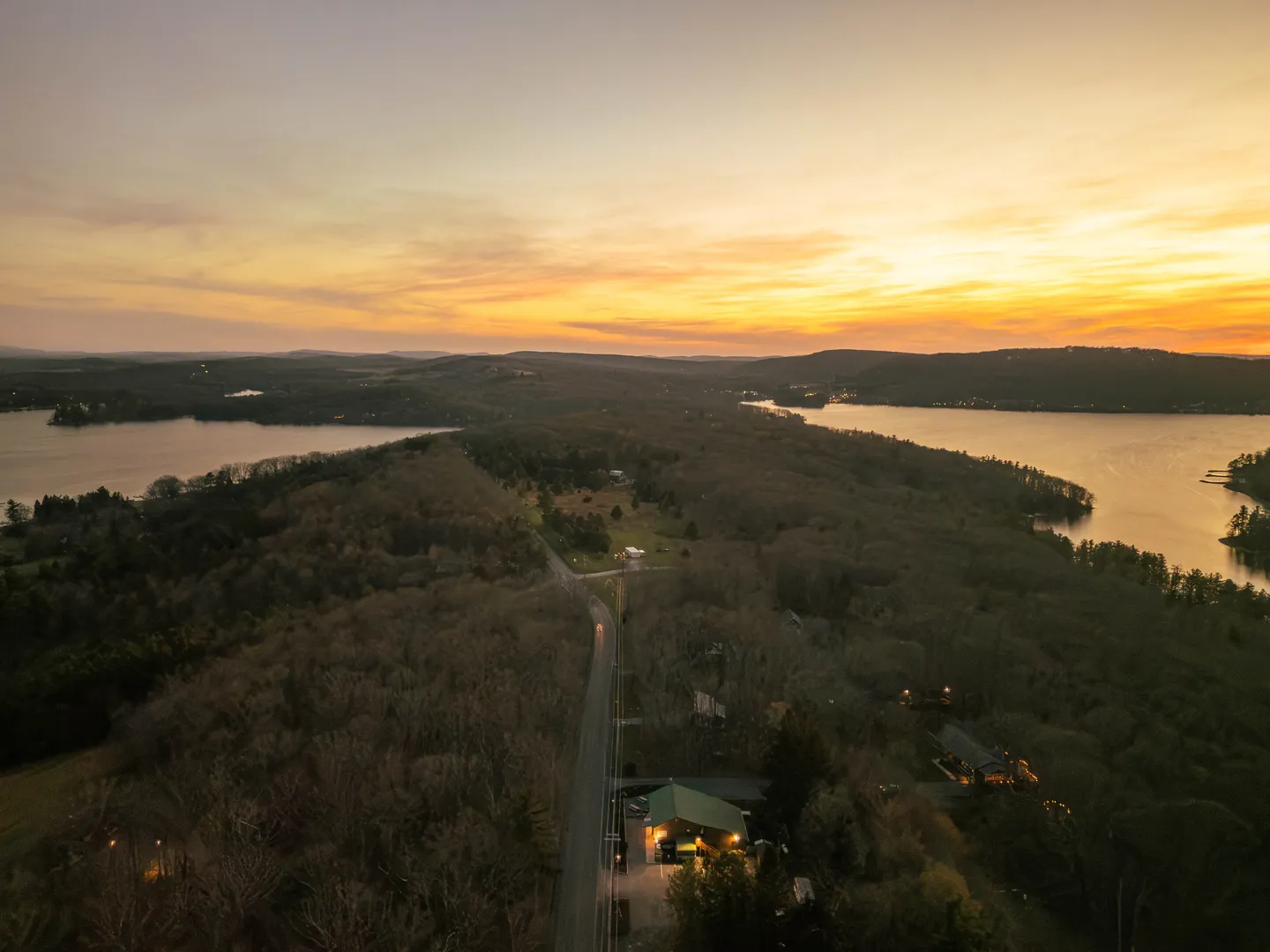 Aerial view of a wooded peninsula bordered by water at dusk, showing a road, scattered buildings, and a glowing sunset sky.