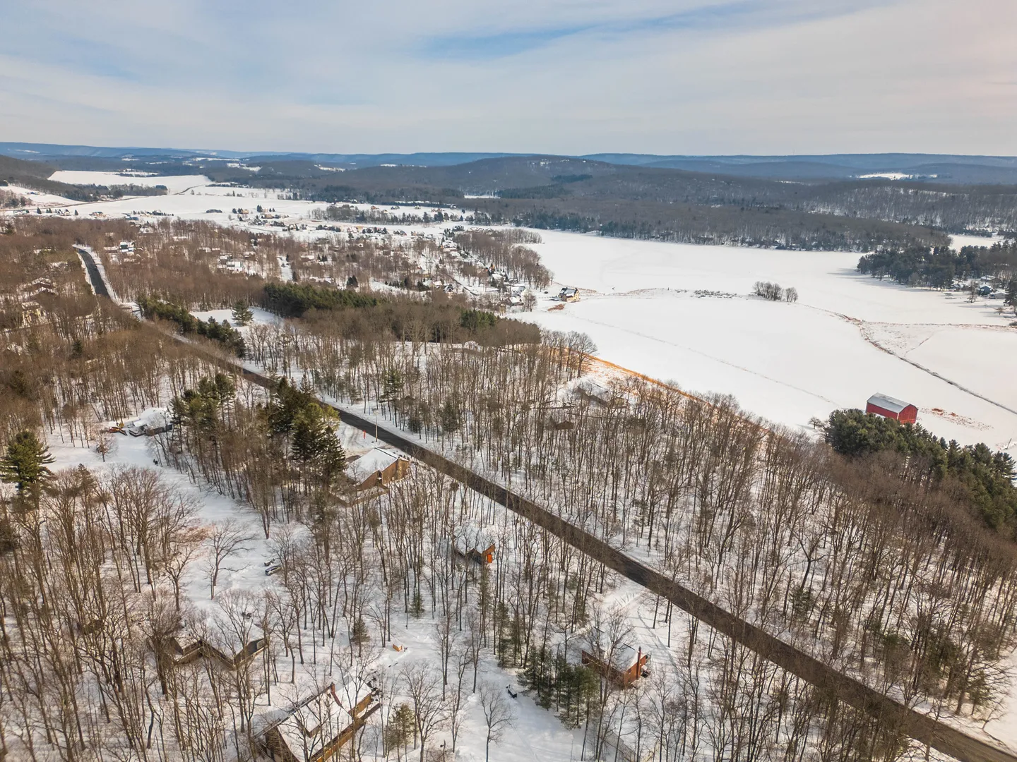 Aerial view of a snow-covered rural landscape featuring a paved road, scattered houses among bare trees, and snowy open fields under a bright sky.
