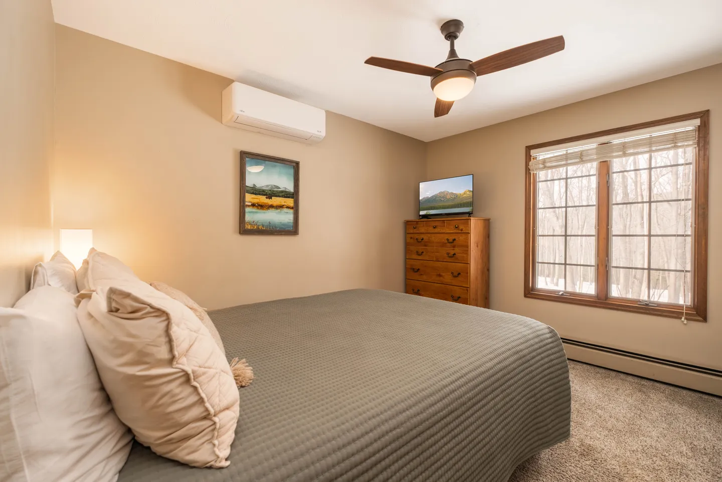 Bedroom with a made bed, wall-mounted air conditioning unit, ceiling fan, wooden dresser with a small TV, and a window.