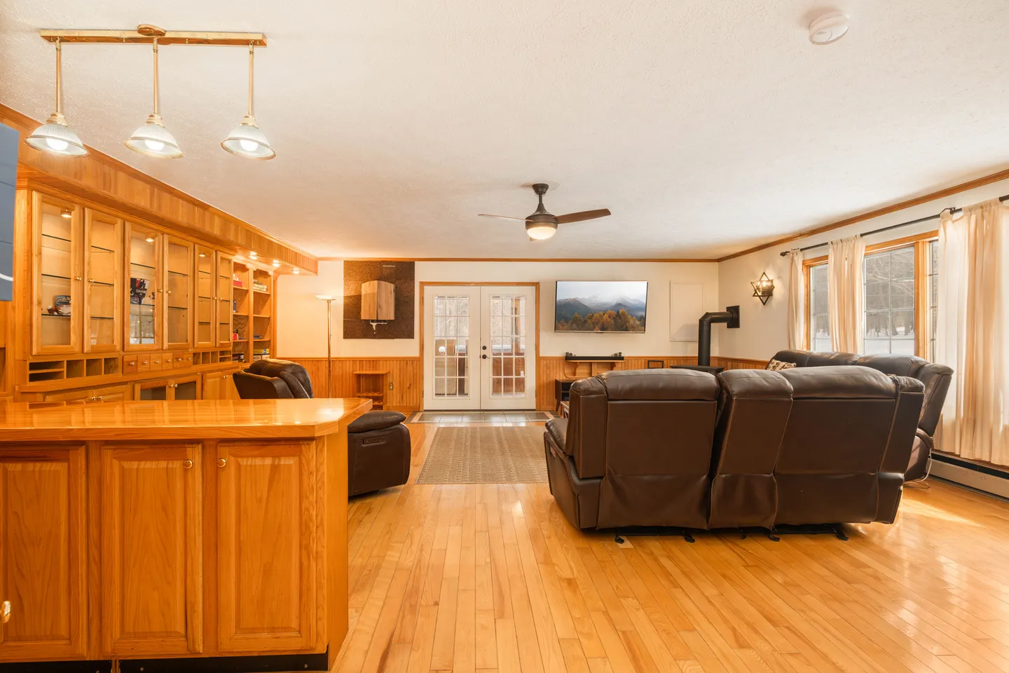 Spacious living room with light wood floors, built-in wooden cabinetry, leather reclining sofas, a mounted TV, and double french doors.