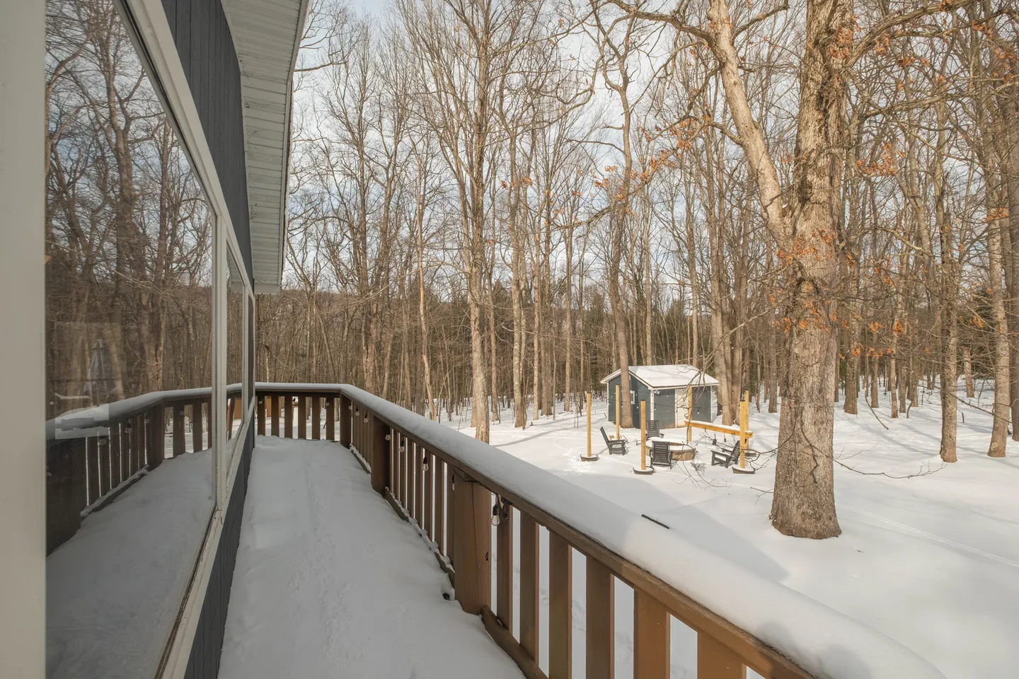 Snow-covered wooden deck railing looking out over a winter forest with a small shed and fire pit area below.