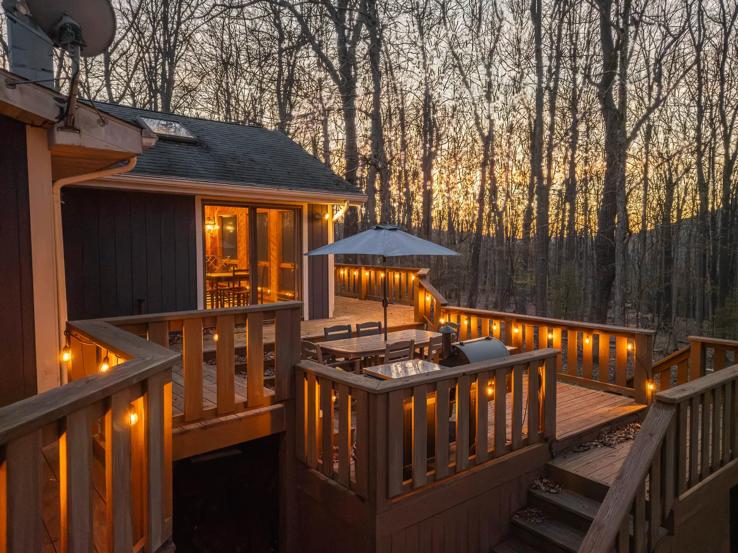 Elevated wooden deck at dusk with outdoor dining table, grill, and string lights overlooking trees.