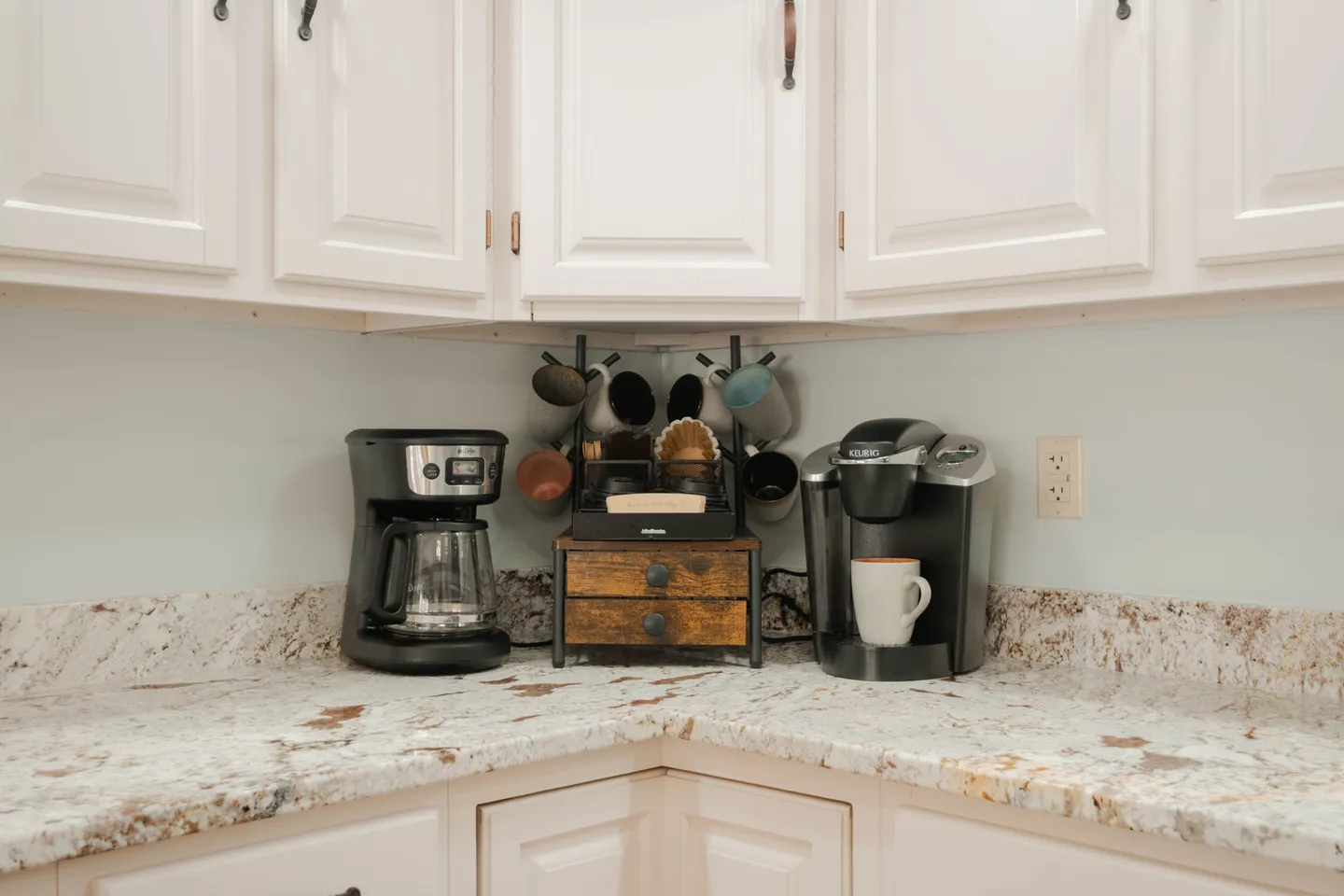 A dedicated coffee station on a speckled countertop beneath white kitchen cabinets, featuring a drip coffee maker, single-serve pod machine, and mug rack.