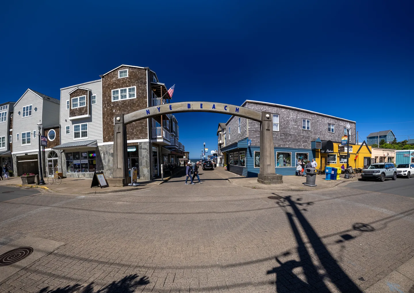 Nye Beach arch sign over a coastal street intersection lined with local shops and multi-story buildings under a clear blue sky.